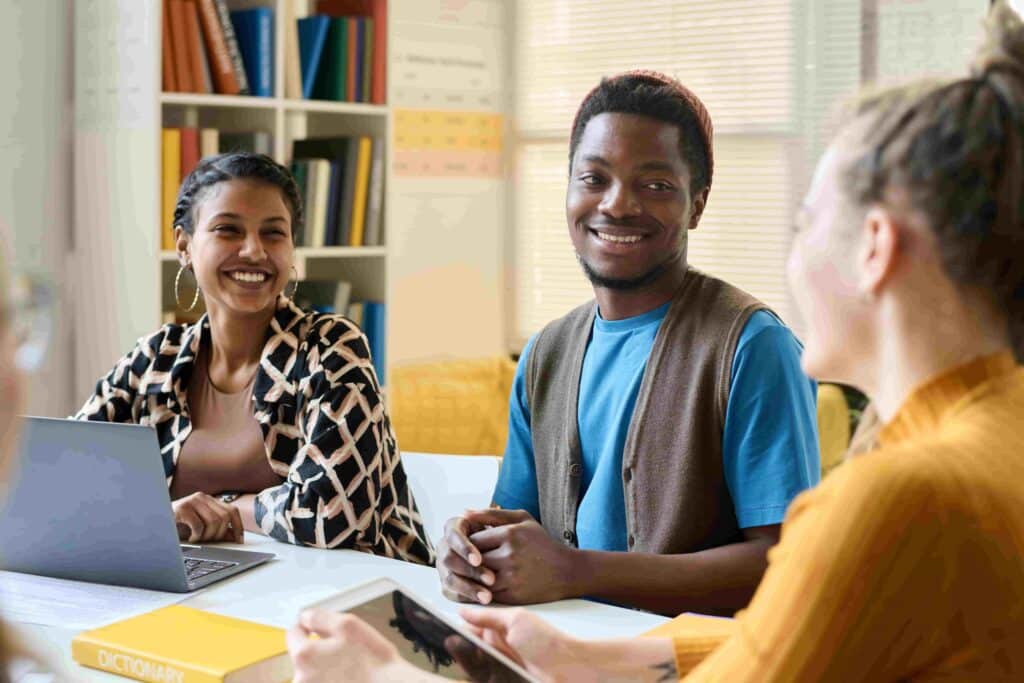 Grupo multiétnico de jovens estudantes alegres rindo enquanto estão sentados à mesa e trabalhando juntos durante as aulas na escola ou faculdade.