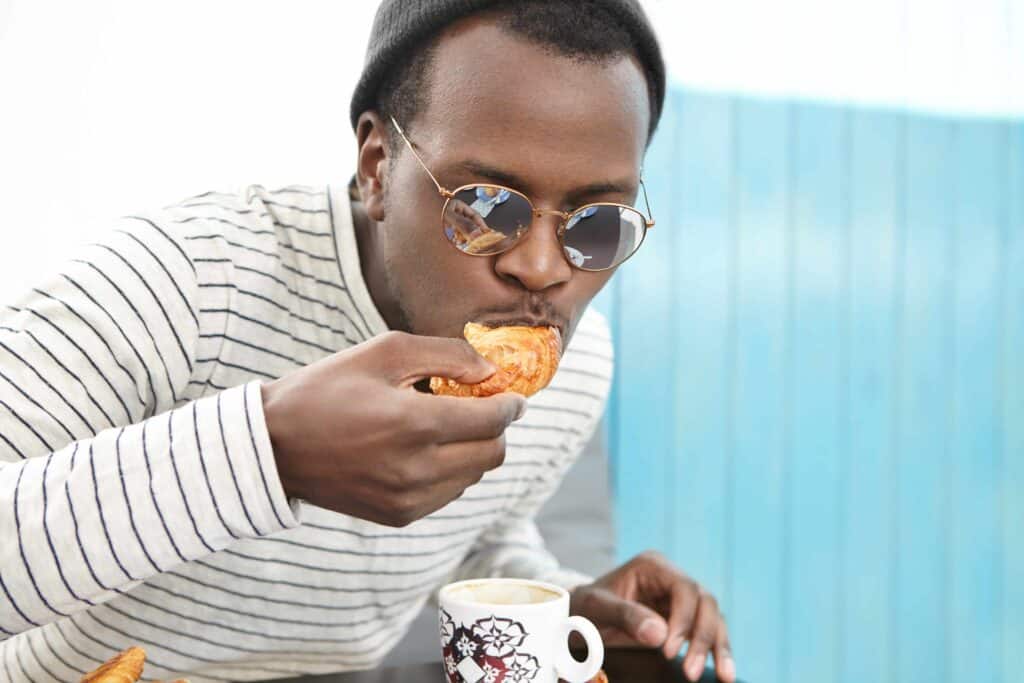 Homem negro de óculos comendo um croissant enquanto toma café. Imagem de fundo azul.