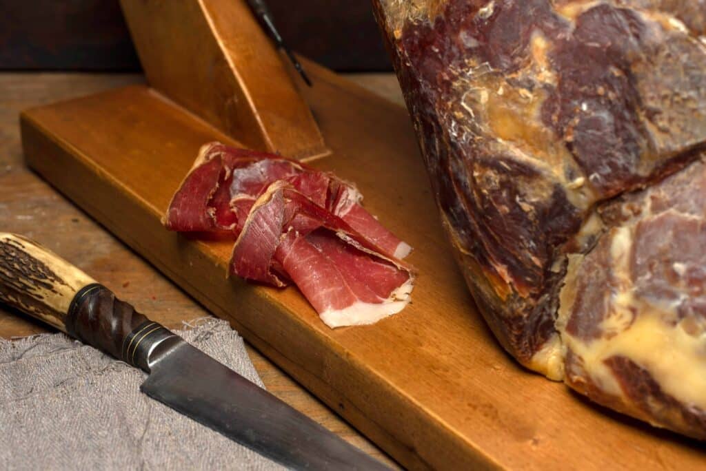 Perna de presunto cru caseiro na tábua de madeira, faca ao lado e fatias de Jamón Espanhol.